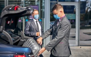 Young man in suit and mask packing businessman luggage into car trunk after his arrival to airport